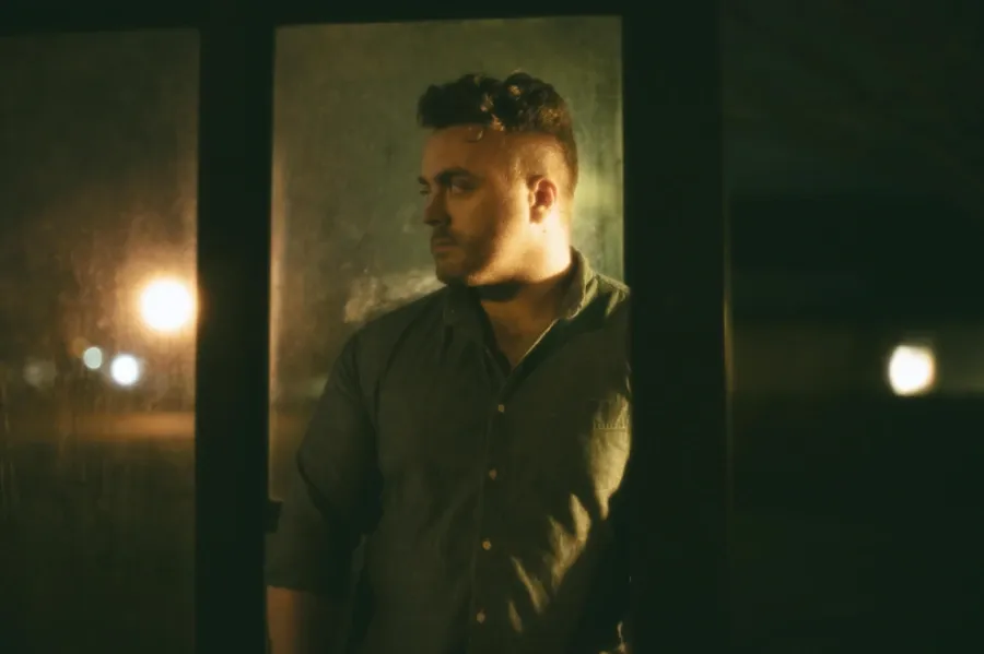 En man står vid ett fönster i ett svagt upplyst rum, med en eftertänksam uttryck, och ser ut genom fönstret. Ljus från utomhuslyktor syns i bakgrunden.