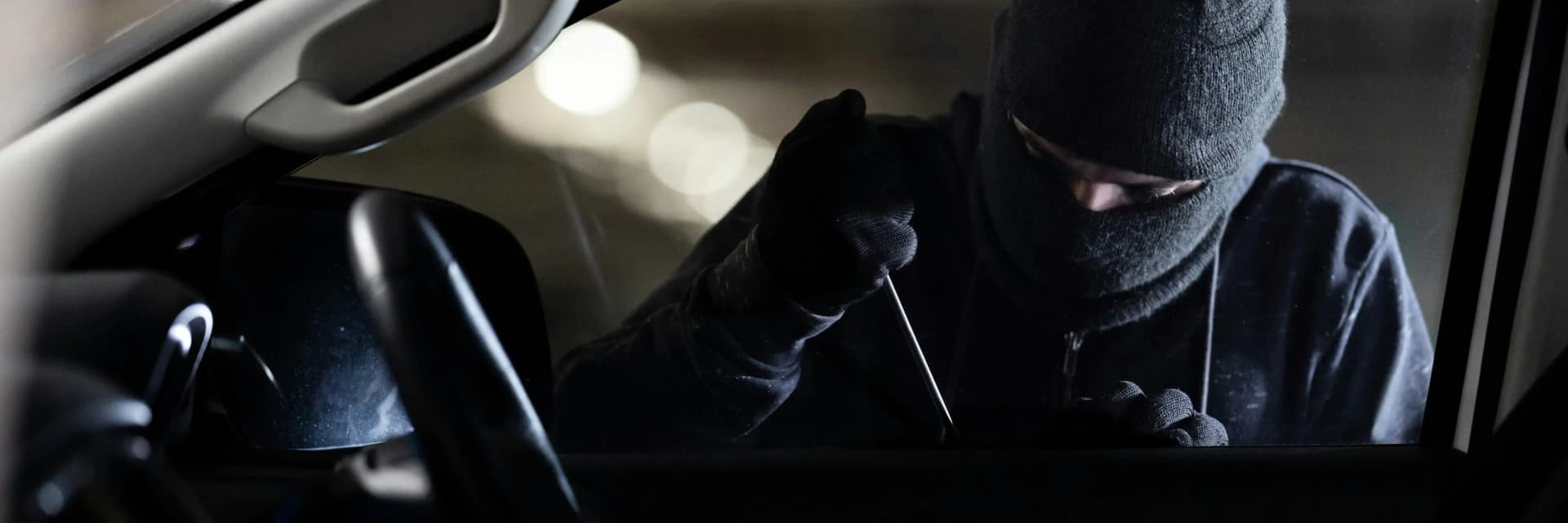 Maskerad person försöker bryta sig in i bil med ett verktyg – illustrerar riskerna med att biluppgifter avslöjar din adress vid camping eller flygplats.