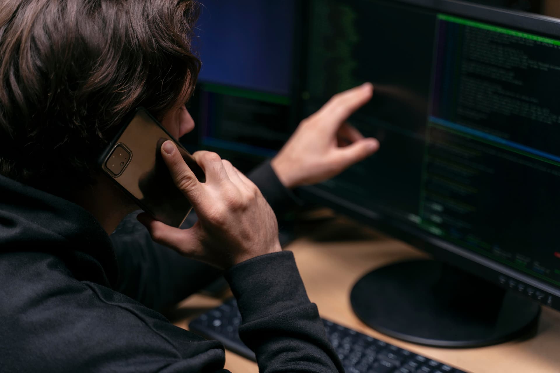 En person sitter vid en dator, pratar i mobiltelefon och pekar mot en skärm med kod och terminalfönster.