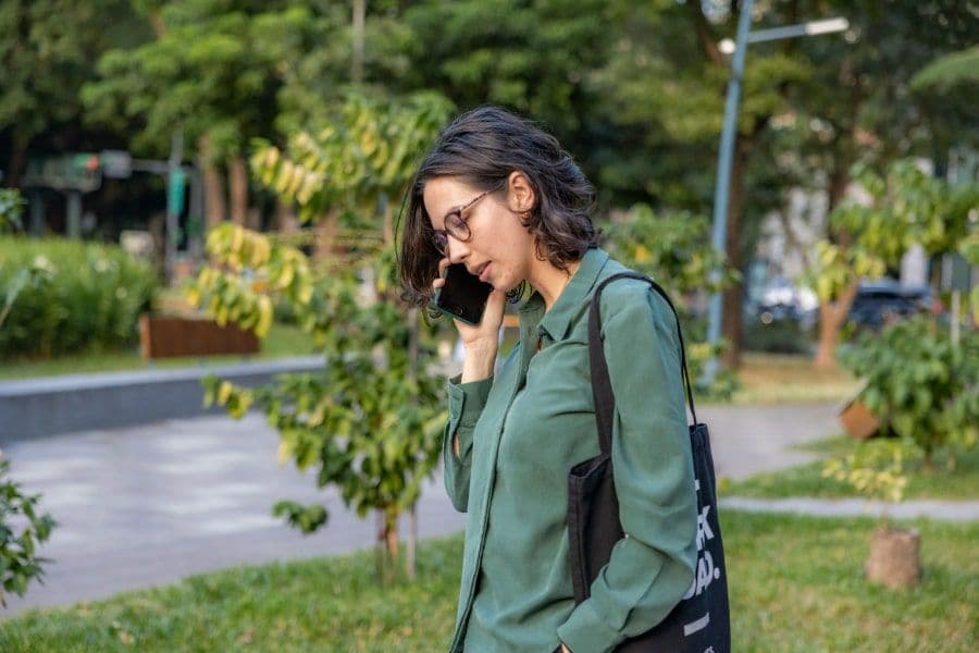 n kvinna med glasögon och grön skjorta pratar i mobiltelefon medan hon promenerar i en park. Hon bär en svart tygväska över axeln och ser allvarlig ut. Grönska och gångvägar syns i bakgrunden.